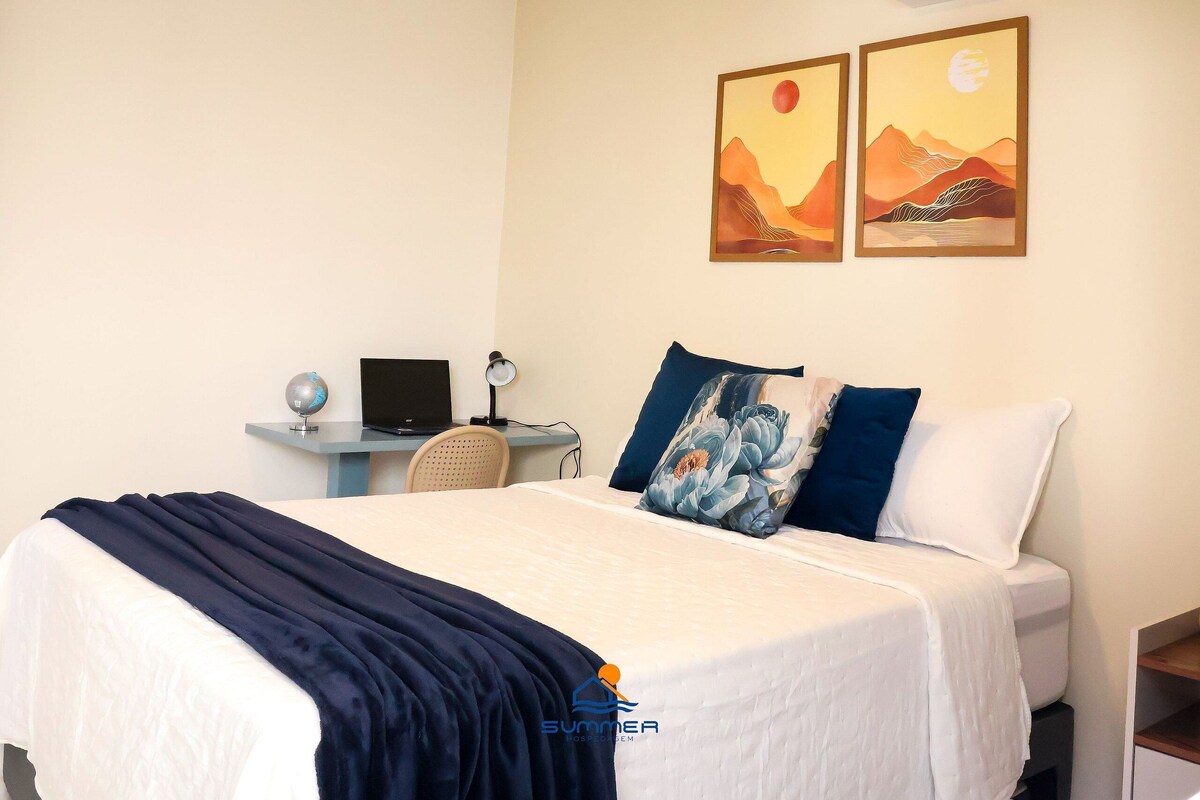 A neatly made bed with a blue blanket, a desk with a laptop, and two wall-mounted paintings.