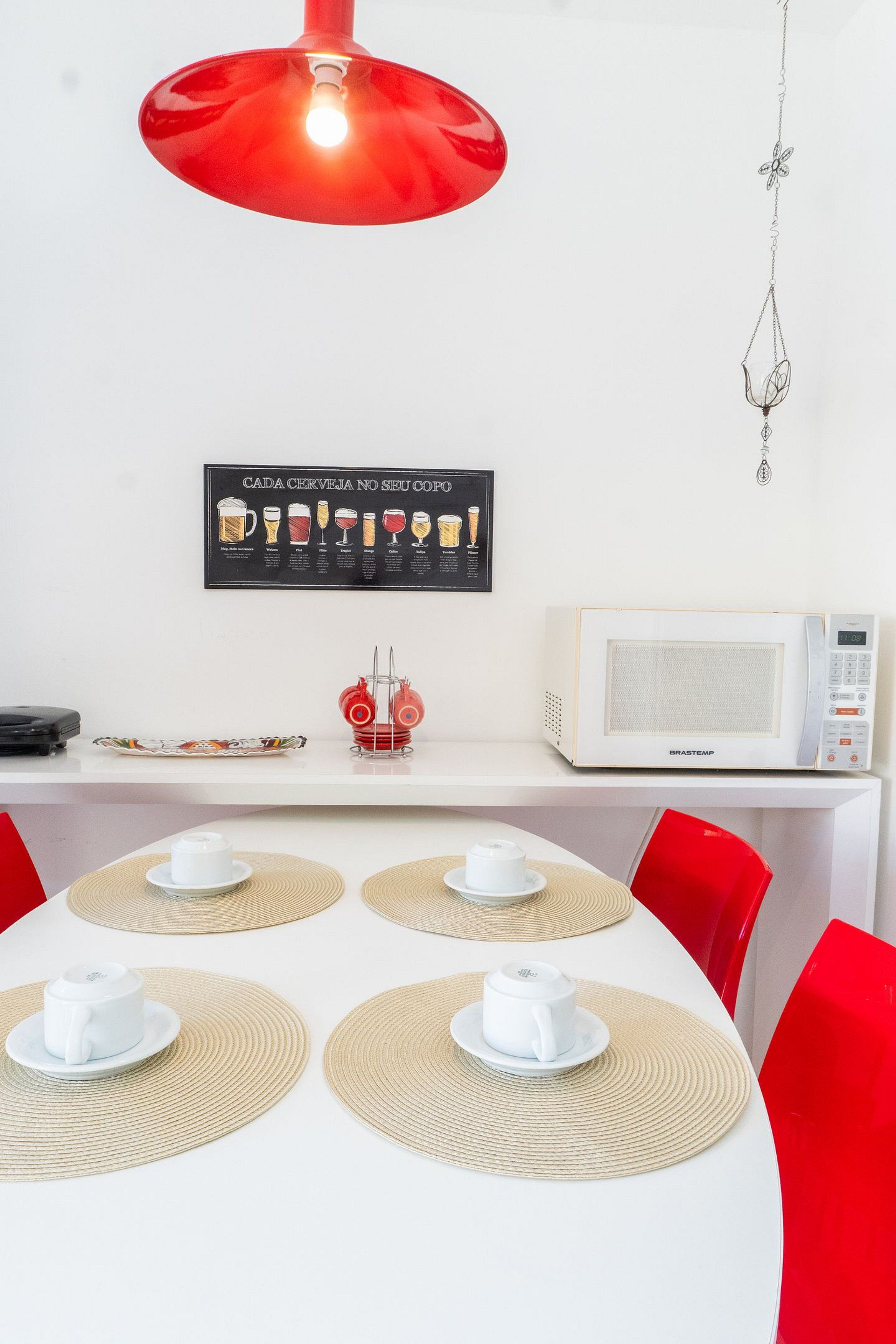 Un comedor con una mesa blanca, sillas rojas, una lámpara colgante roja, un microondas BraTaste y un tablero de menú en la pared.
