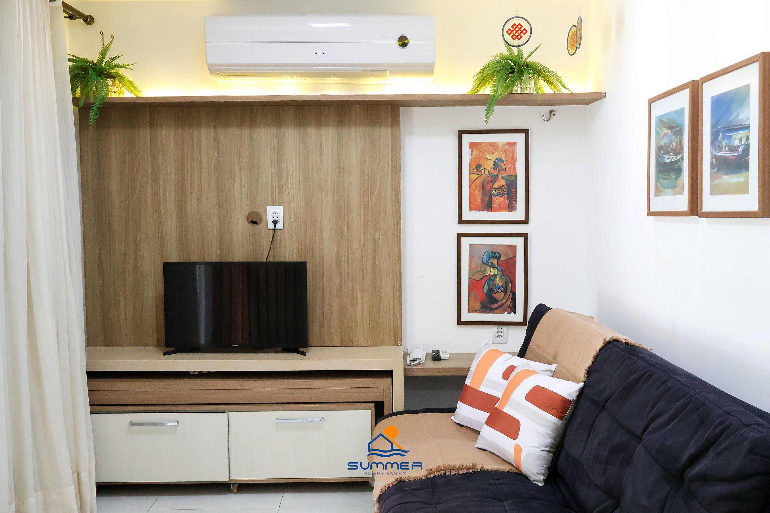 A modern living room with a flat-screen TV, wooden panel walls, a sofa with patterned cushions, and framed artwork on the wall.