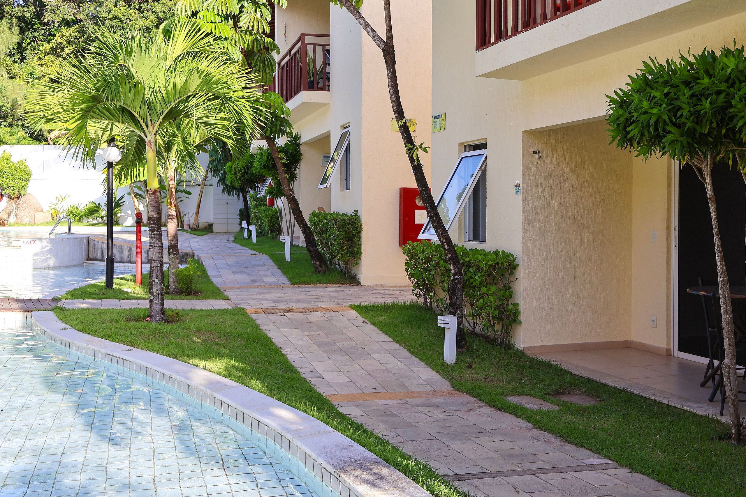 Un área junto a la piscina con un camino de baldosas, palmeras y un edificio con balcones y una puerta.