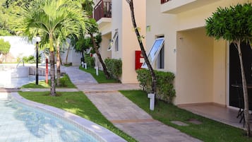 Un área junto a la piscina con un camino de baldosas, palmeras y un edificio con balcones y una puerta.