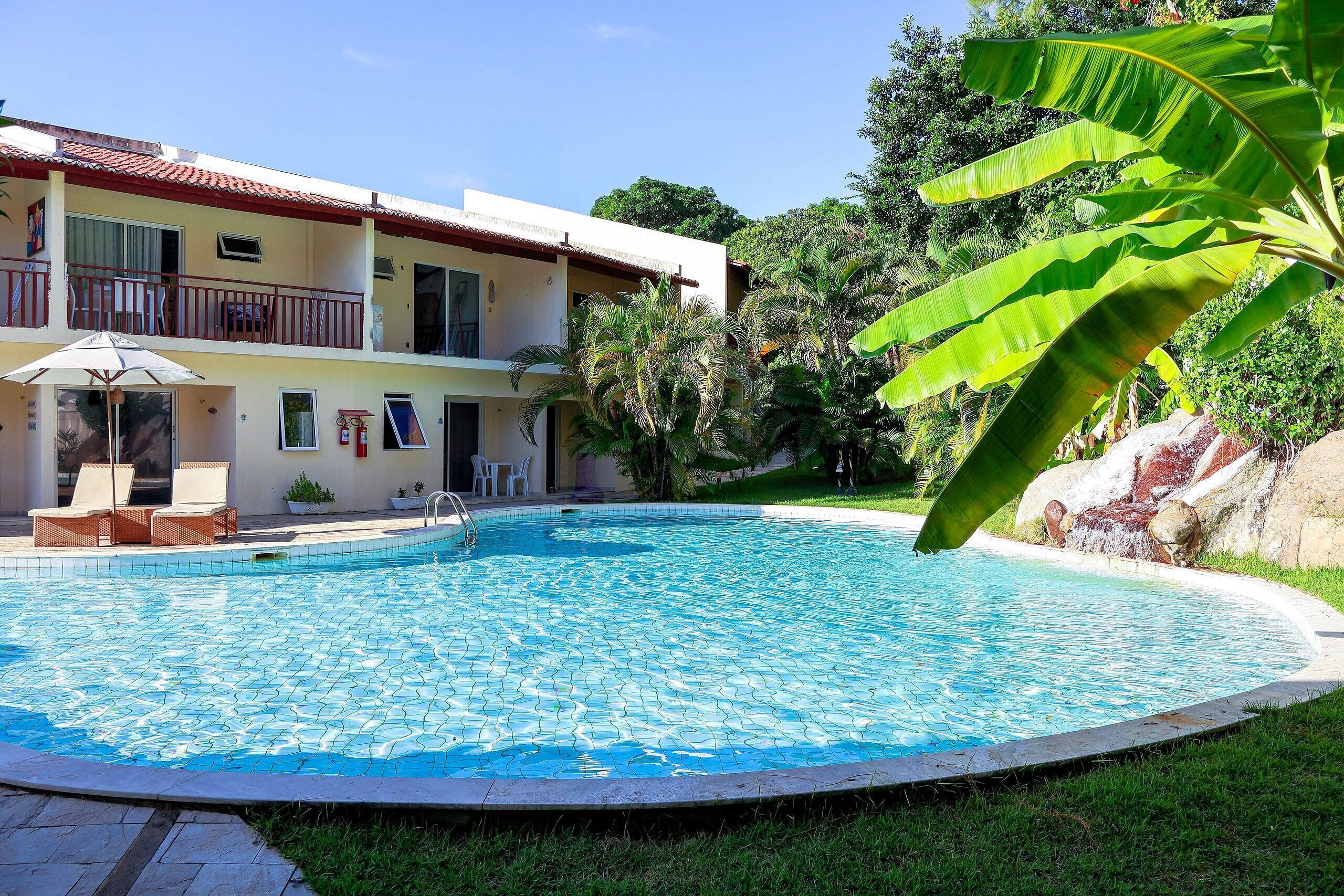 A swimming pool with a tiled deck, a two-story building with balconies, and lush greenery.