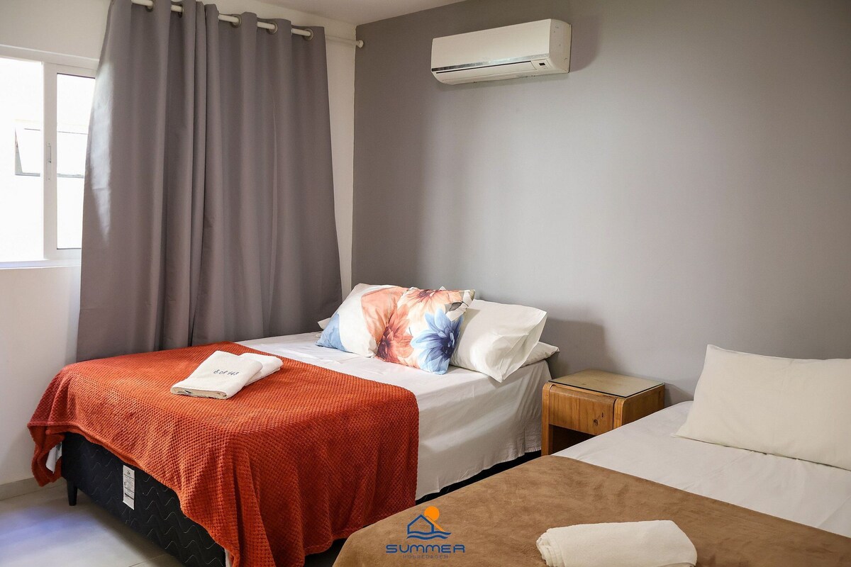 A hotel room with a bed, a nightstand, an air conditioning unit, and a window with curtains.