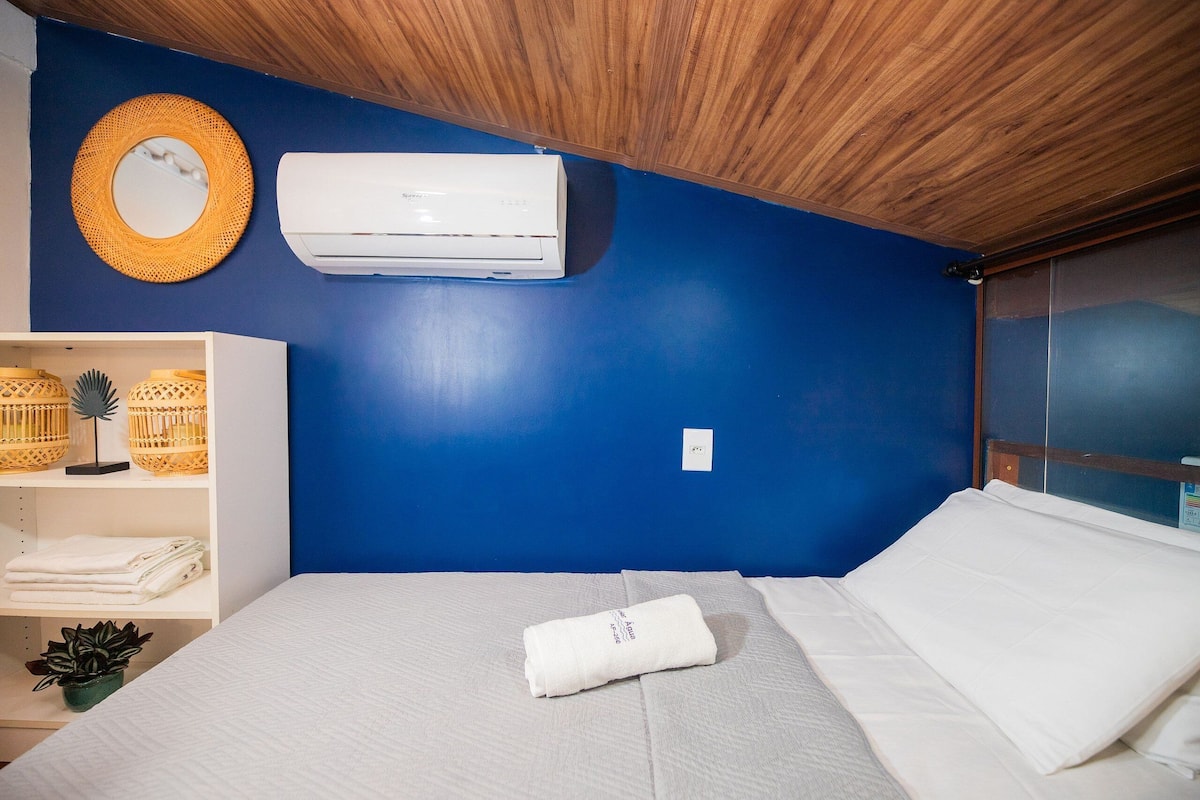 Una habitación con pared azul, aire acondicionado blanco, techo de madera, cama con ropa blanca, estante blanco con cestas y un espejo.