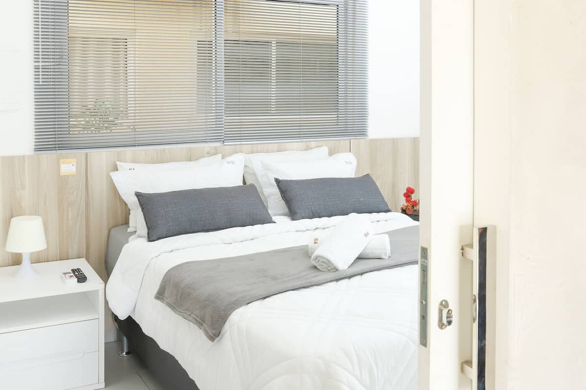 Una cama bien hecha con una colcha blanca y almohadas grises, una lámpara de noche y una ventana con persianas.