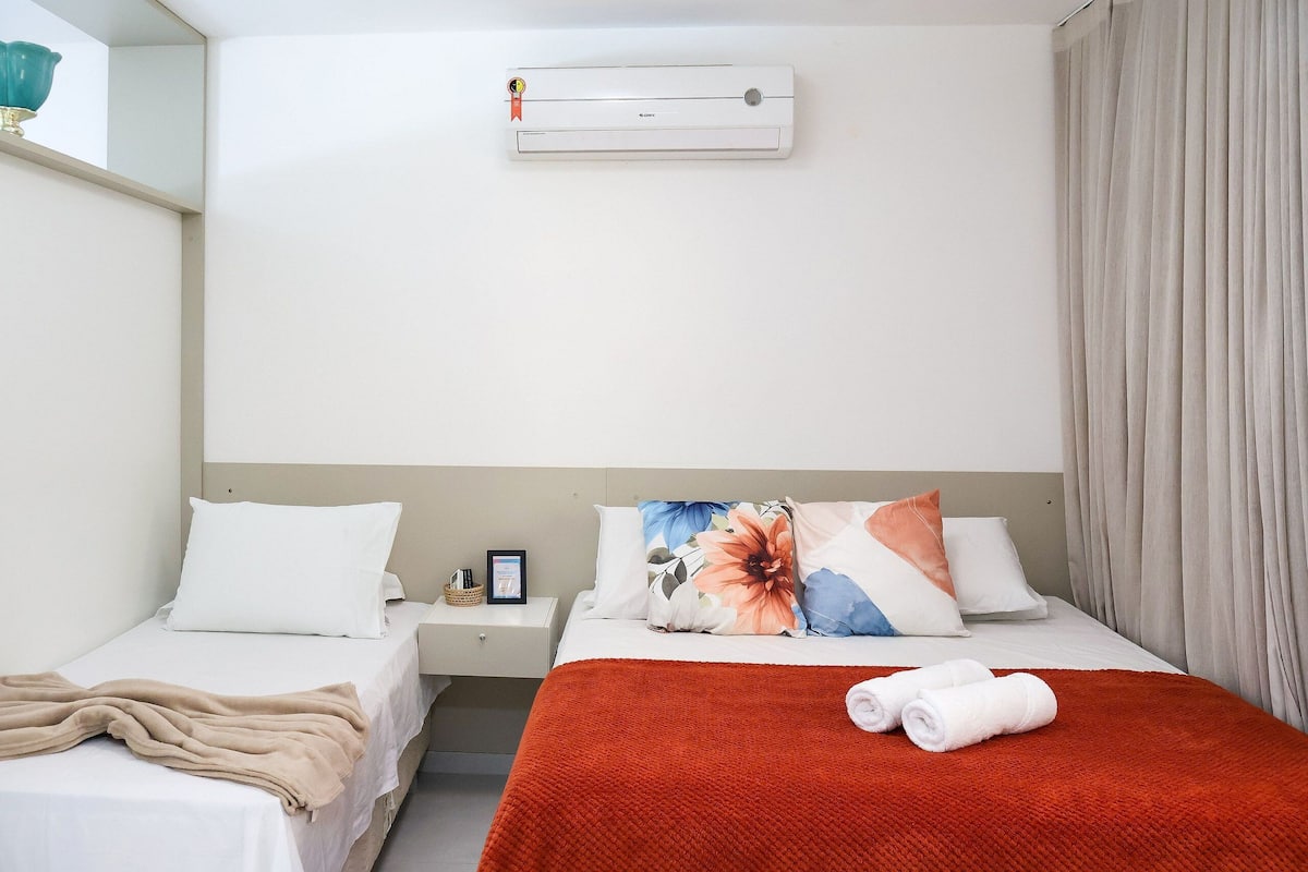 Una habitación de hotel con dos camas, una colcha roja sobre la cama más grande, ropa de cama blanca, una mesita de noche con una lámpara y un equipo de aire acondicionado en la pared.