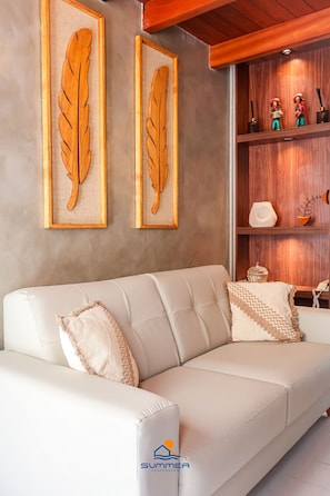 A white leather sofa with decorative pillows, framed leaf artwork on the wall, and a wooden shelf with figurines.