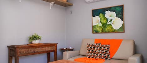 A living room with a sofa, a coffee table, a wooden shelf with decorative items, a painting, and an air conditioning unit.