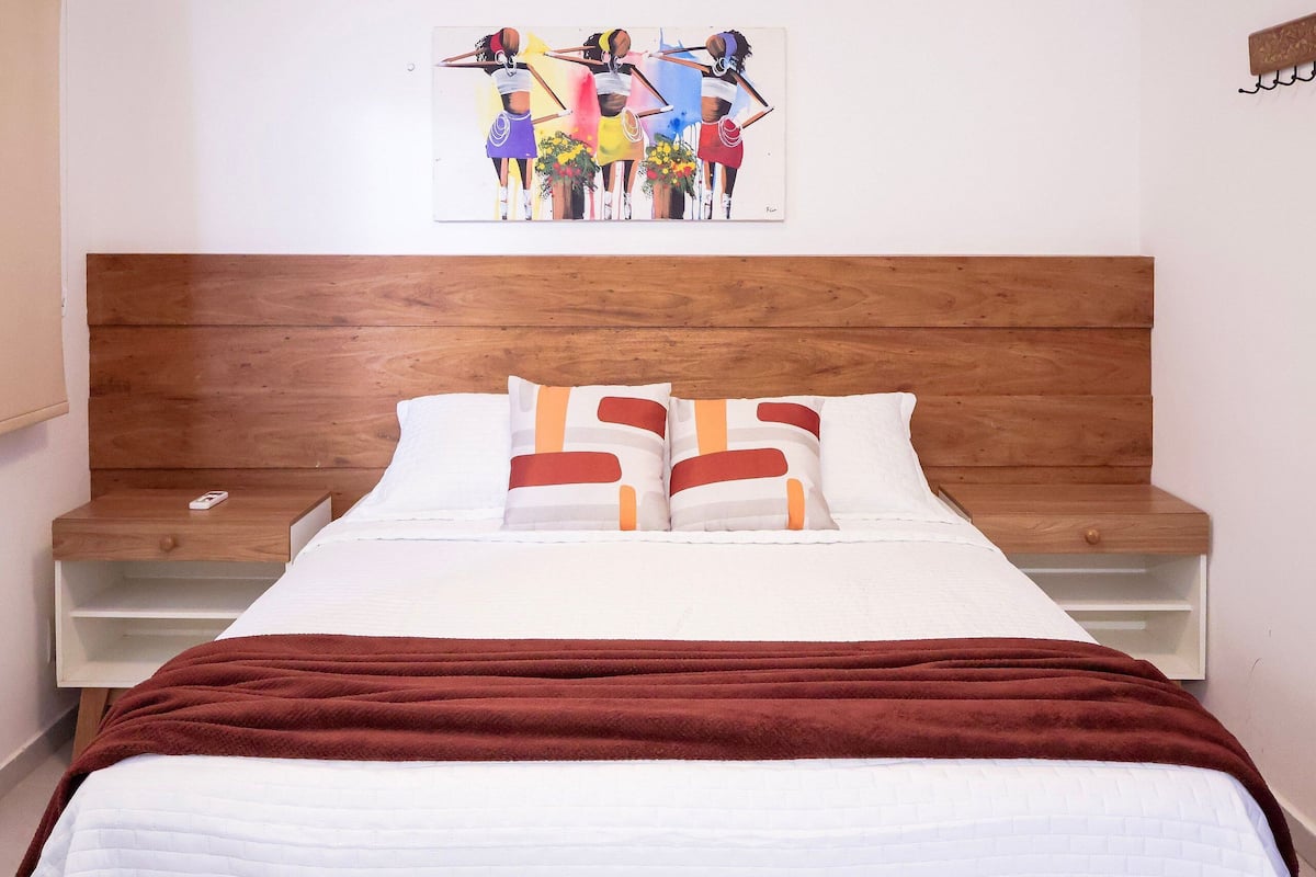 Una cama con cabecera de madera, ropa de cama blanca y una manta marrón. Hay una mesita de noche con un teléfono y una obra de arte colgada en la pared sobre la cama.