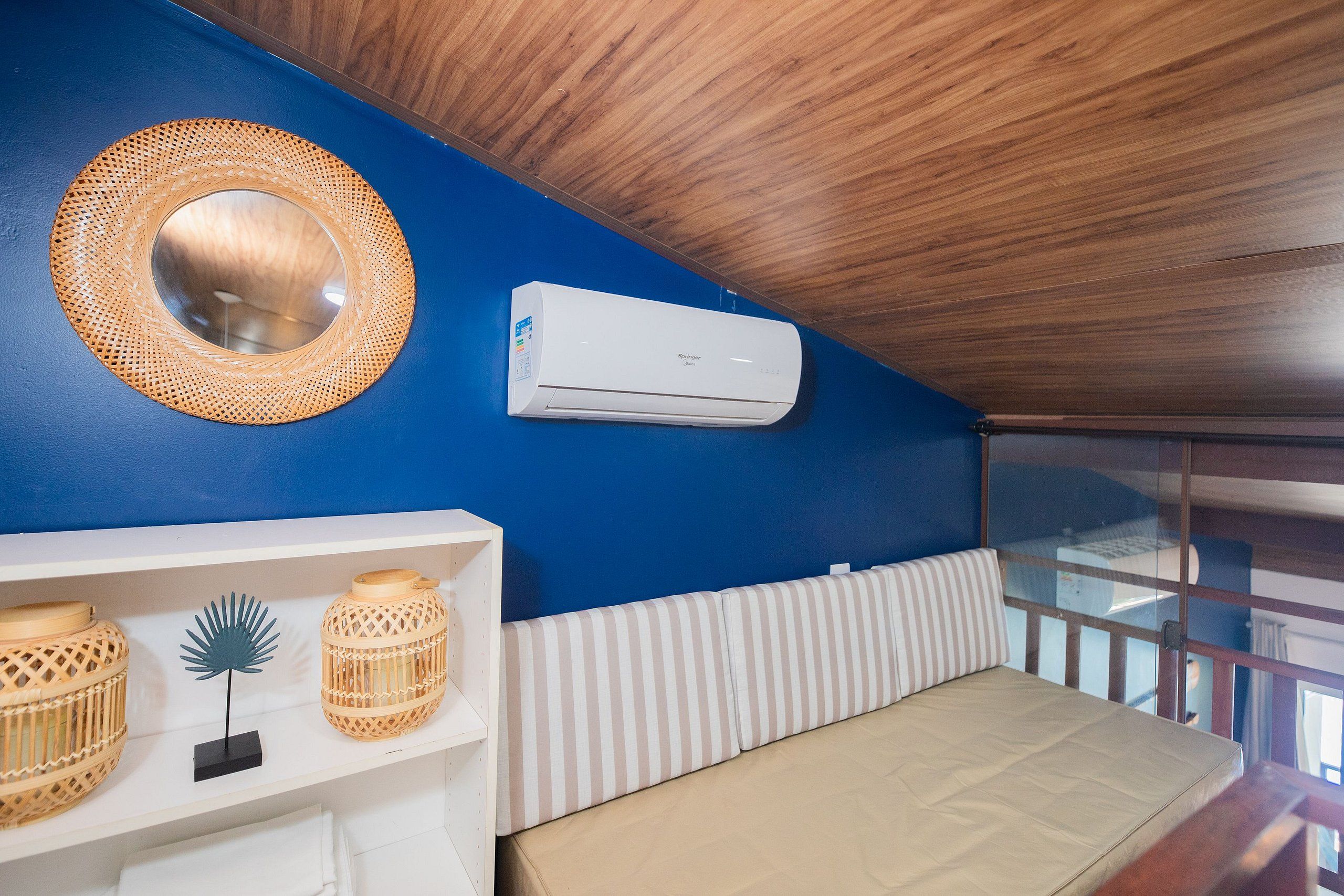 A room with a blue wall, a round woven mirror, a white air conditioning unit, a white shelf with wicker baskets, and a striped bench.