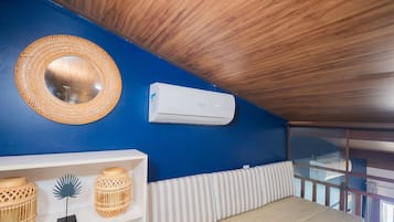 A room with a blue wall, a round woven mirror, a white air conditioning unit, a white shelf with wicker baskets, and a striped bench.