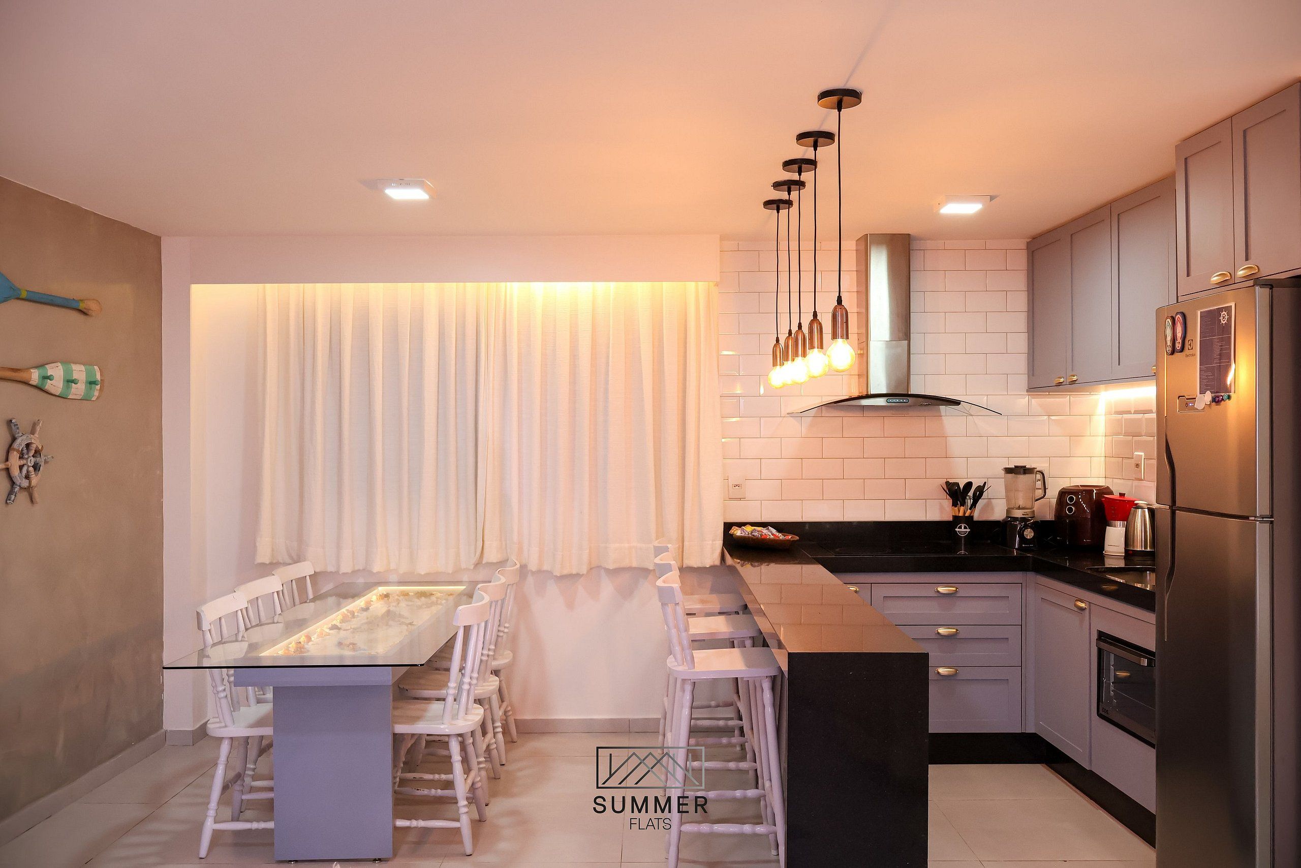 Uma cozinha moderna com área de jantar, destacando uma ilha central, luminárias pendentes e eletrodomésticos de aço inoxidável.