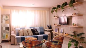 Ein Wohnzimmer mit einer Couch, Sesseln, einem Couchtisch, einem Fernseher, Pflanzenregalen und einer Vitrine mit Flaschen.