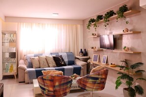 A living room with a sofa, armchairs, a coffee table, a TV, shelves with plants, and a glass cabinet with bottles. - Solar Água Pipa- Summer Pipa (Tibau do Sul)