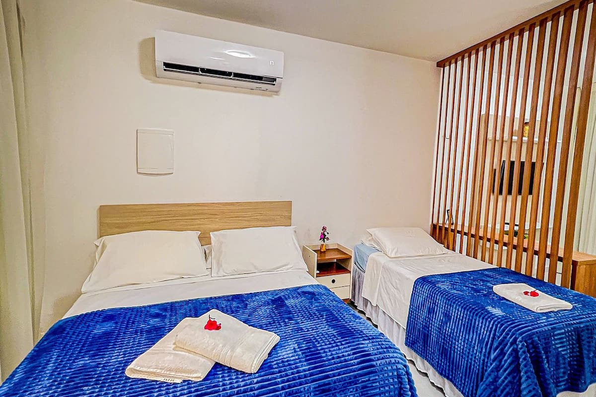 Dos camas con ropa de cama azul, una lámpara en el techo, un equipo de aire acondicionado y un separador de madera con listones.