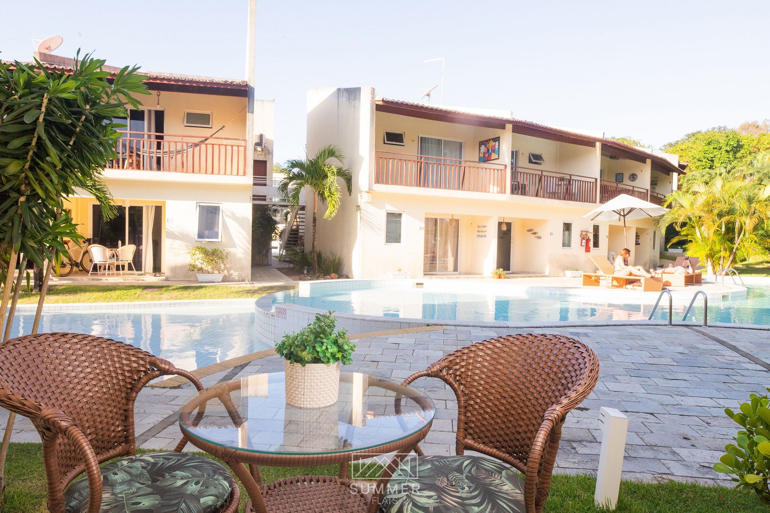 Zona junto a la piscina con mobiliario de mimbre, una mesa de vidrio y un edificio con balcones y una piscina.