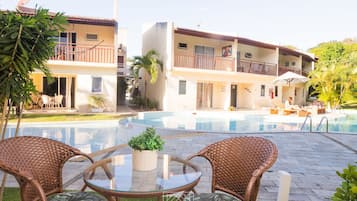 Zona junto a la piscina con mobiliario de mimbre, una mesa de vidrio y un edificio con balcones y una piscina.