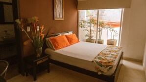 A bedroom with a bed, a nightstand, a chair, a window with a view of plants, and a framed picture on the wall. - Toukan Hostel (Pereira)