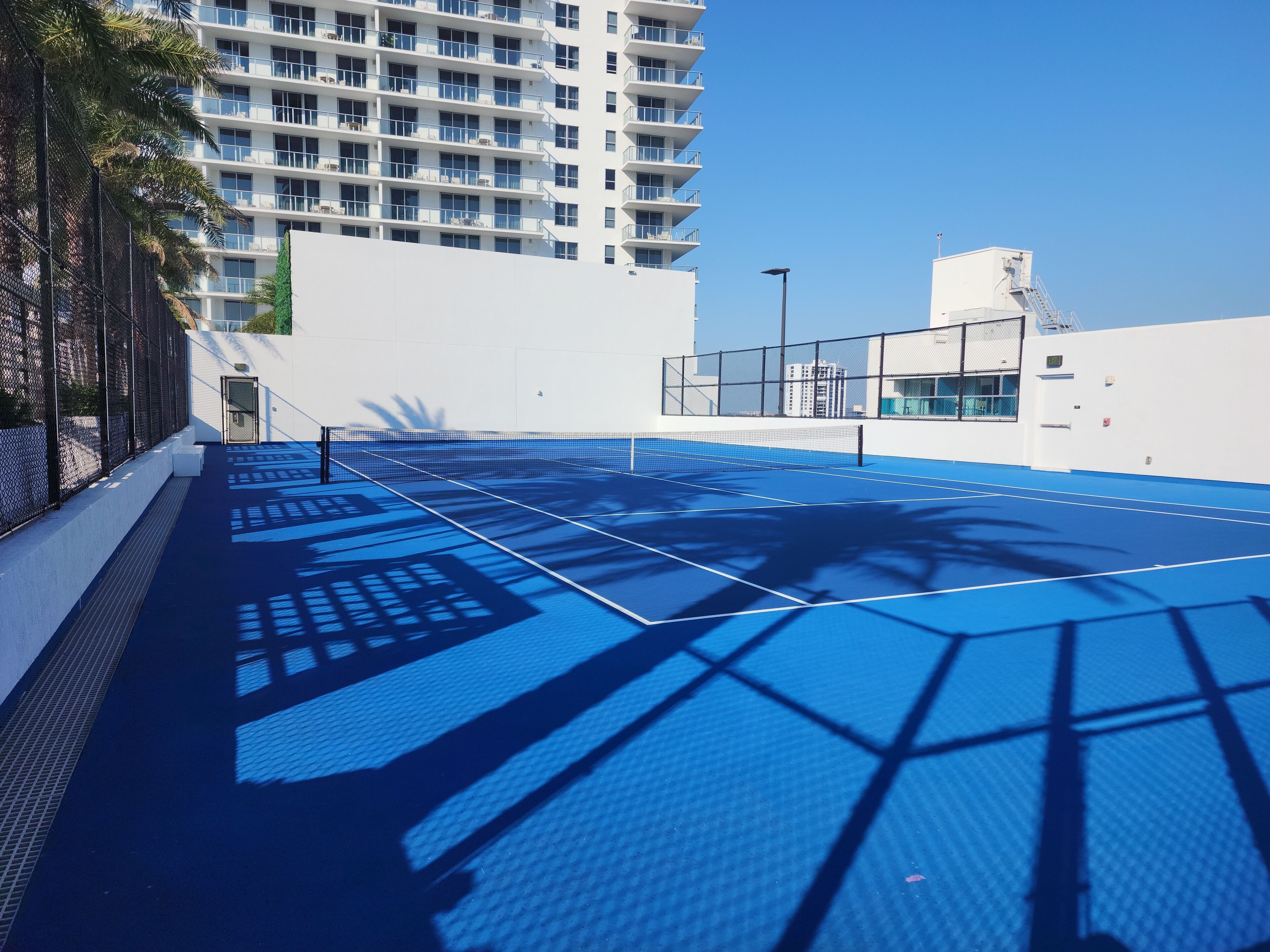 Uma quadra de tênis ao ar livre com superfície azul, cercada por paredes brancas e um edifício alto ao fundo.