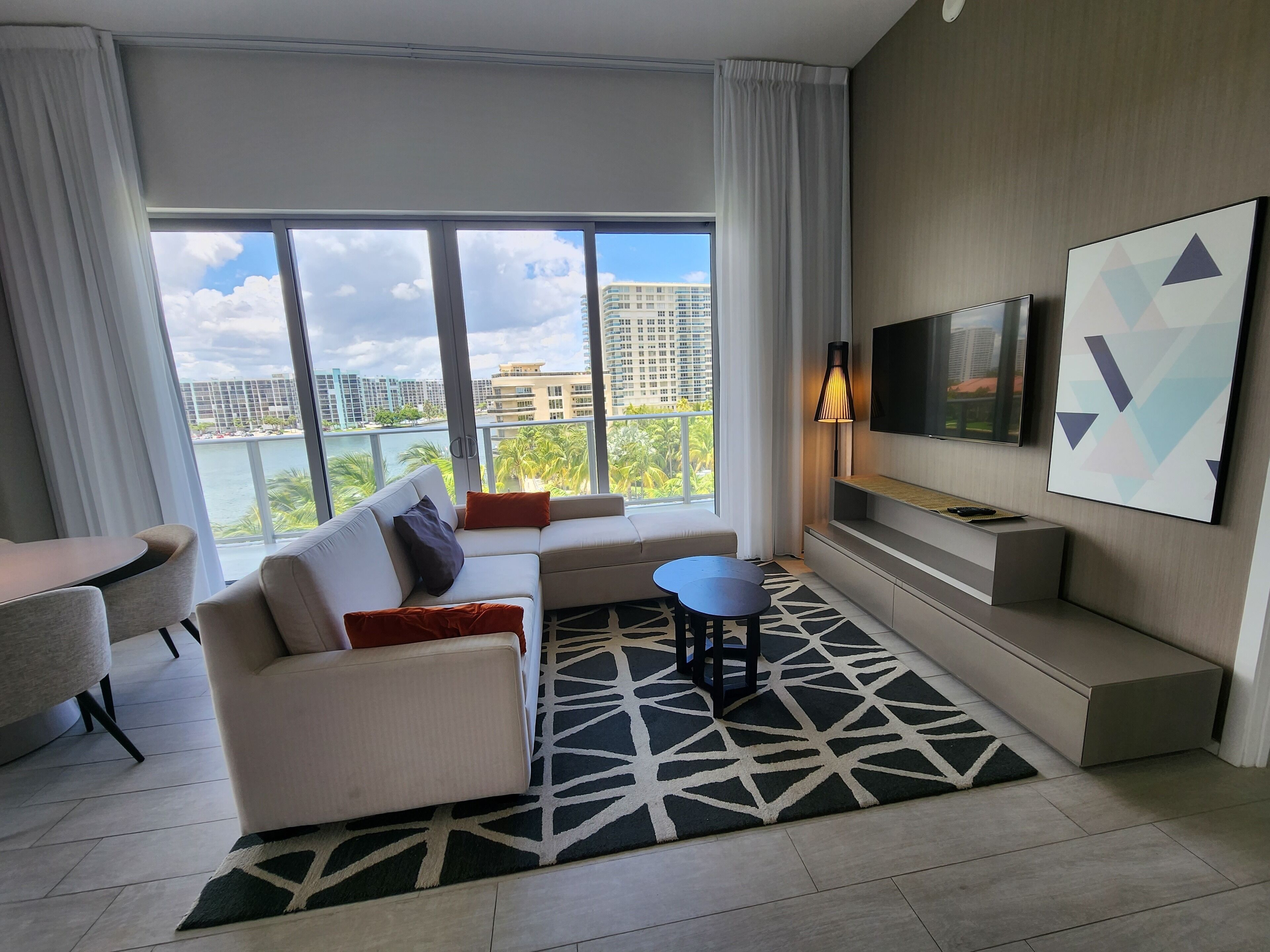 A modern living room with a sofa, a small table, a TV mounted on the wall, and a large window with a city view.