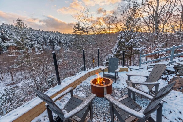 Una terraza cubierta de nieve con un hoyo de fuego y sillas Adirondack.
