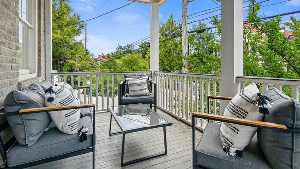 A covered porch with a glass table and two gray sofas with striped pillows, overlooking a green landscape with trees and power lines. - Spring St - Downtown - Walk Everywhere! (Charleston)