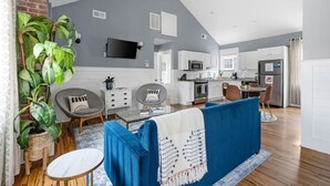 A modern living room with a blue sofa, a coffee table, and a TV mounted on the wall. There is a kitchen area with white cabinets and a refrigerator. - Spring St - Downtown - Walk Everywhere! (Charleston)