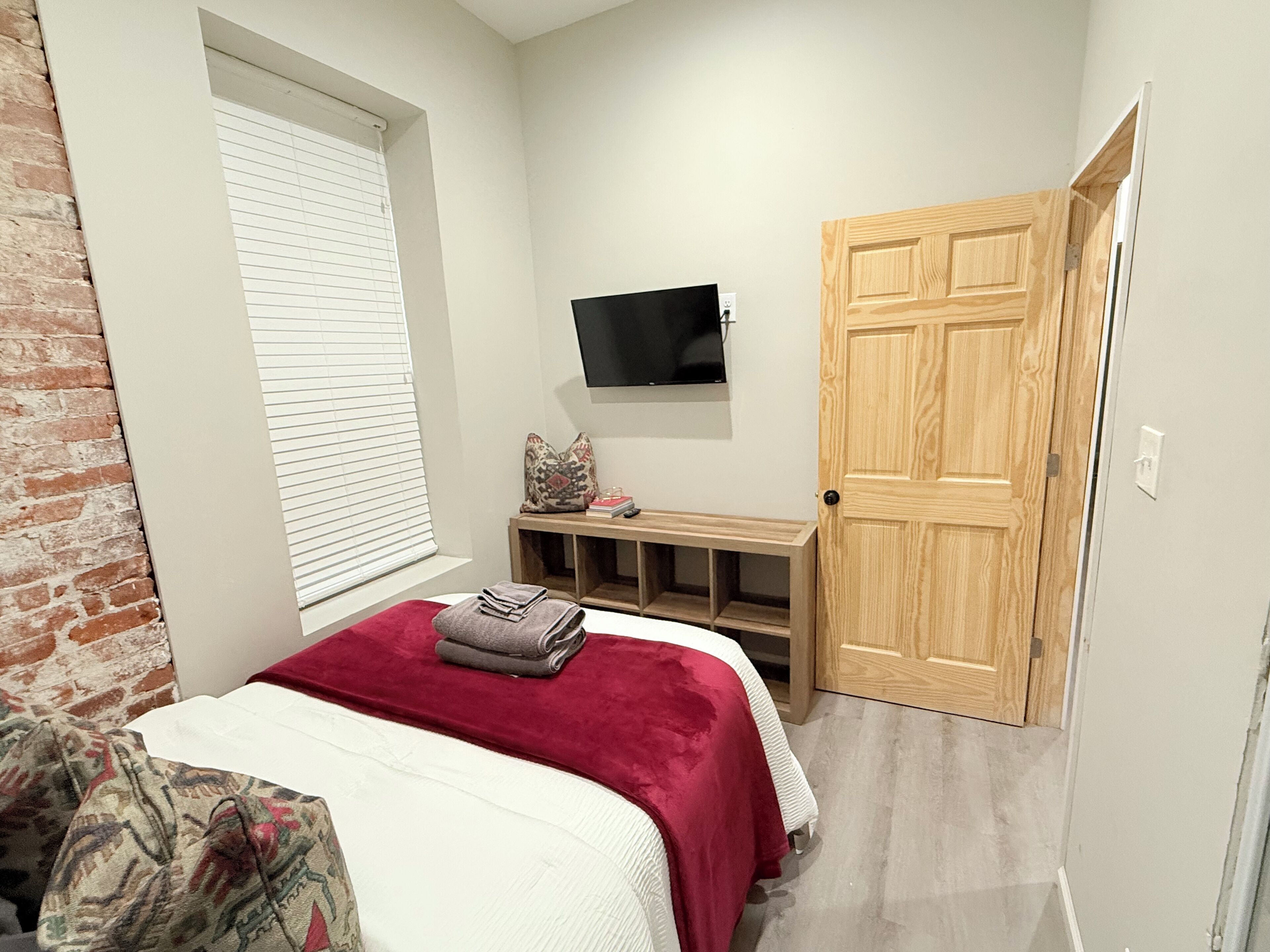 A bedroom with a bed, a TV mounted on the wall, a wooden door, and a window with blinds.