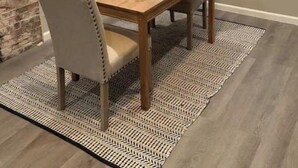 A dining area with a wooden table and two chairs, a patterned rug, and a brick wall.