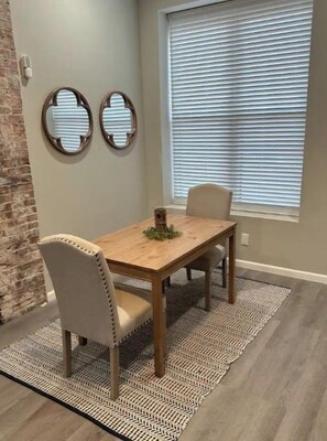 A dining area with a wooden table and two chairs, a patterned rug, and a brick wall.