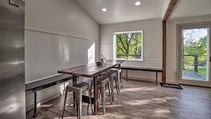 Una cucina moderna con un lungo tavolo di legno e sgabelli da bar, un'ampia finestra con vista su un'area verde, e un tappetino d'ingresso.