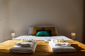 Ein ordentlich gemachtes Bett mit einer gelben Tagesdecke, weißen Bettwäsche und einem grünen Kissen. Zwei Nachttischlampen und Handtücher liegen auf dem Bett.