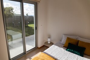 Ein Schlafzimmer mit Bett, einem kleinen Tisch und Blick auf einen Garten durch eine Schiebetür.