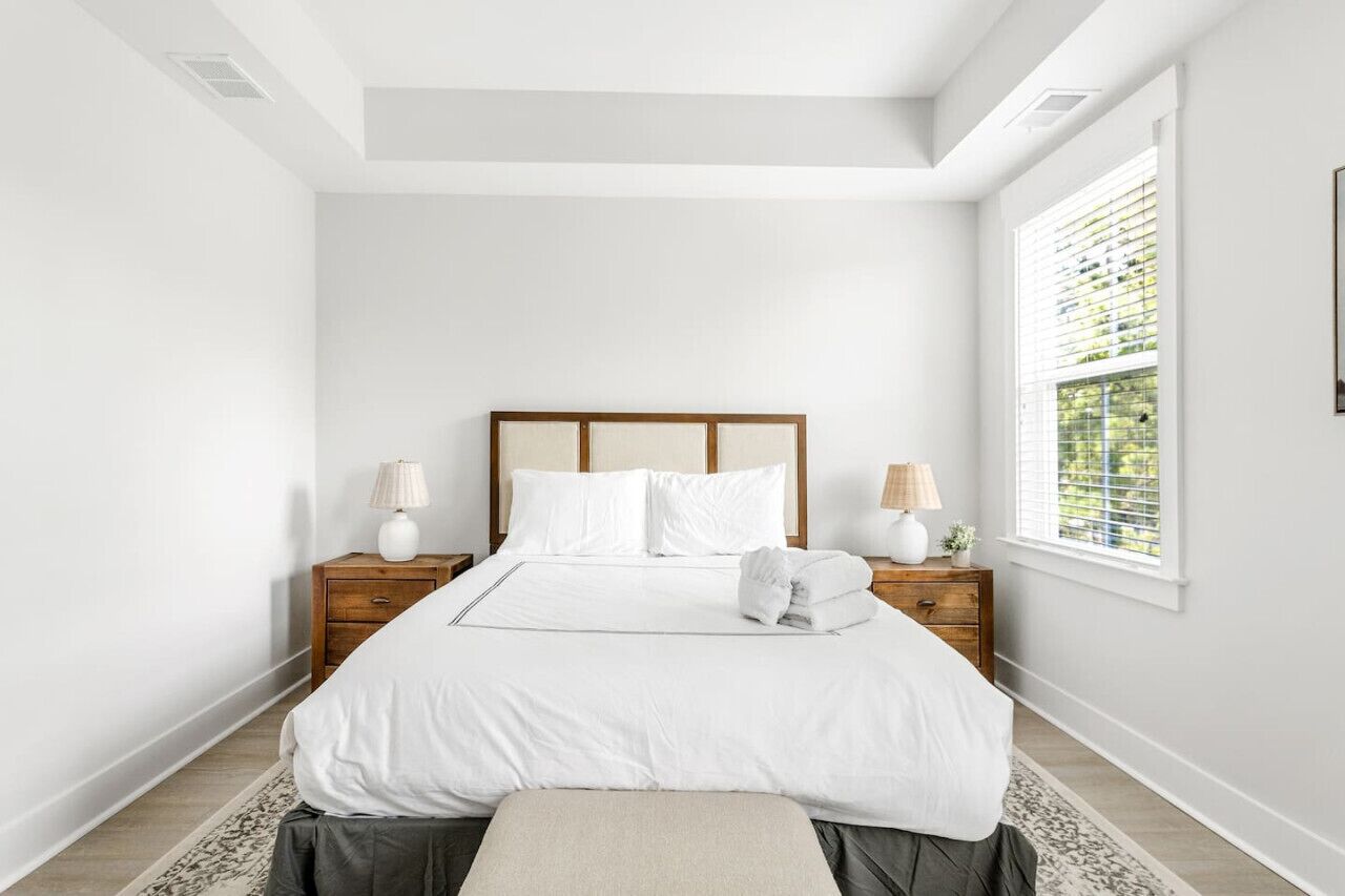 Une chambre à coucher bien rangée, avec un grand lit, deux tables de chevet avec des lampes, une fenêtre avec des stores et une petite plante sur la table de chevet.