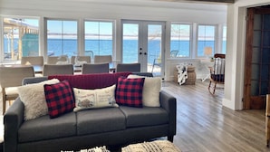 Ein gemütliches Wohnzimmer mit grauem Sofa, mehreren Sesseln und Blick auf das Wasser durch große Fenster.