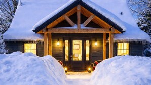 Eine Blockhütte mit schneebedecktem Dach, einer Veranda mit zwei Laternen und einer Auffahrt zum Eingang.
