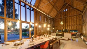 A spacious dining area with a long table set for a meal, large windows, and a cozy interior with wooden walls. - Grand Suffolk Barn Conversion (Suffolk)