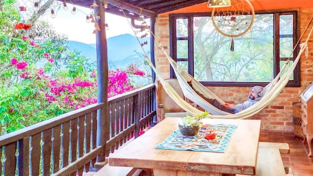 Un hamac sur une terrasse en bois, avec une table, un luminaire suspendu et une vue sur des fleurs roses et de la verdure.