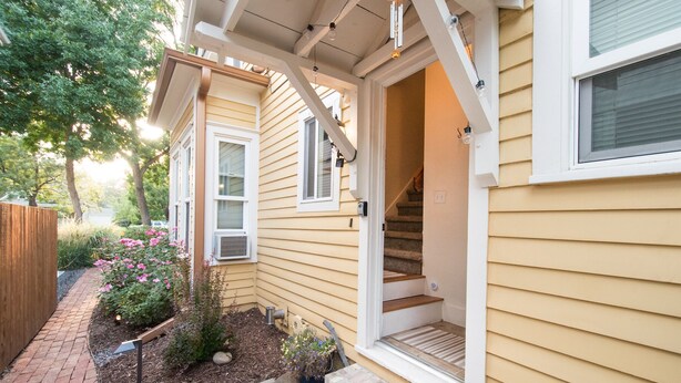 Une maison jaune avec une terrasse en bois, un chemin en briques et un petit jardin parsemé de fleurs roses.