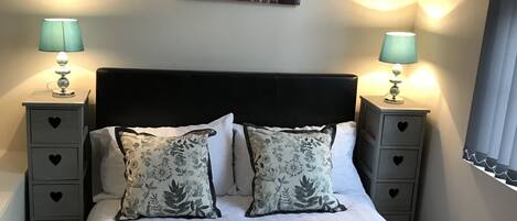 A neatly made bed with floral pillows, two bedside tables with lamps, a wall-mounted artwork, and a window with blinds.