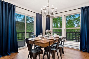 Comedor con mesa de madera dispuesta para cuatro personas, sillas oscuras, ventanas amplias con cortinas azules y una araña.
