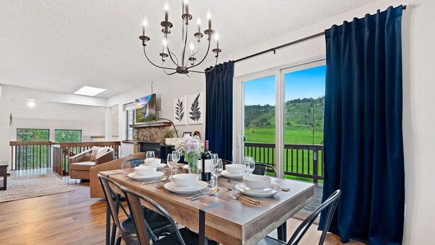 Un comedor con una mesa de madera dispuesta para cuatro, una araña y vistas a un paisaje verde a través de amplias ventanas.