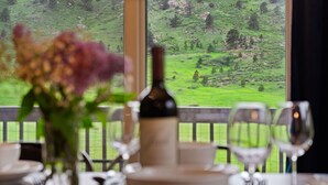 Una mesa de comedor con una botella de vino, un florero con flores y copas de vino, frente a un paisaje pintoresco que se ve a través de una ventana.