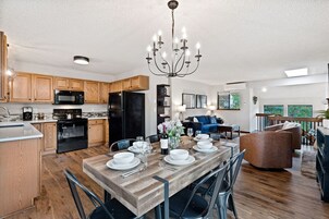 A modern kitchen with wooden cabinets, a dining area with a wooden table set for six, and a living area with a blue sofa and a brown armchair.