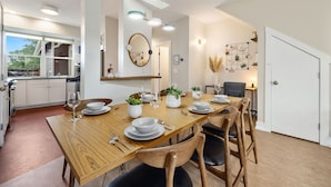A modern dining area with a wooden table set for four, surrounded by wooden chairs. A kitchen with white cabinets and a dishwasher is visible to the left. - Mid-century Mod Retreat Near Old Town – Free Bikes (Fort Collins)
