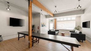 A spacious room with a long table, two sofas, a flat-screen TV, and a large window providing natural light.