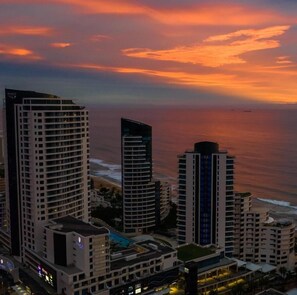 Un paisaje urbano costero con rascacielos, incluyendo una estructura curva distintiva, y vista del océano al atardecer.