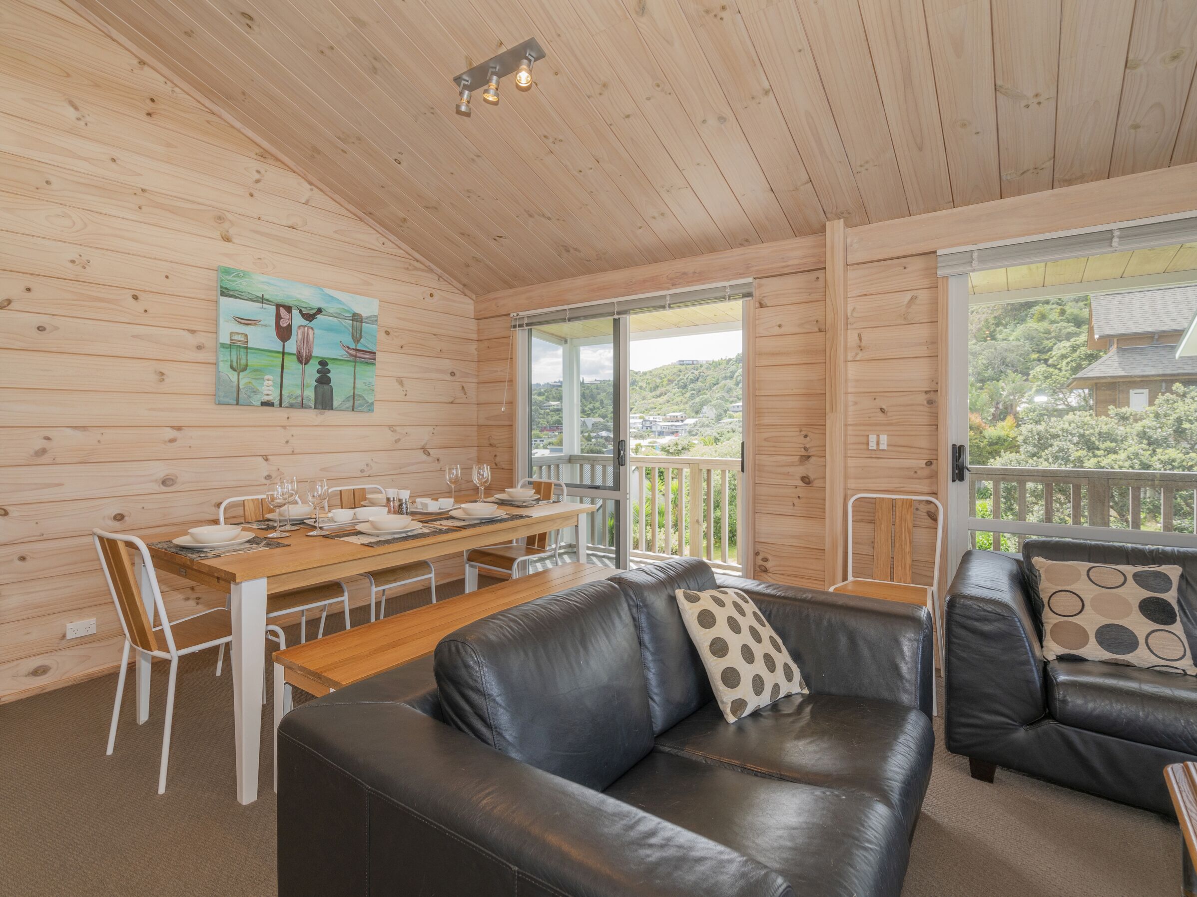 A wooden cabin interior with a dining table set, a leather sofa, and a painting on the wall.