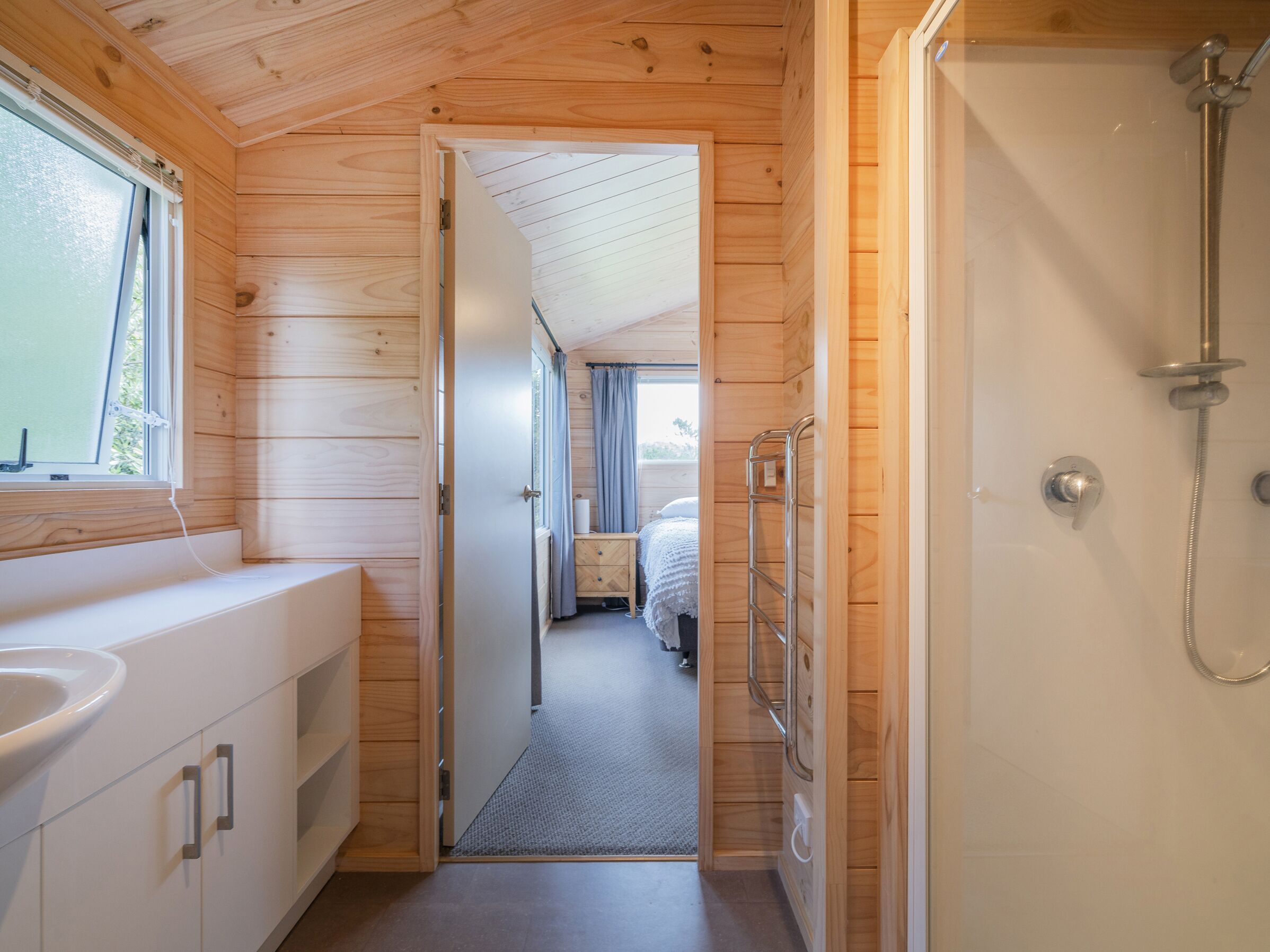 A wooden cabin interior with a bathroom featuring a shower, a sink, and a bed with a window.