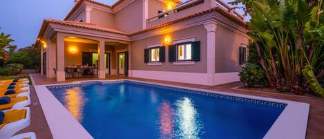 Una casa de dos pisos con piscina, mobiliario de exterior y plantas tropicales.